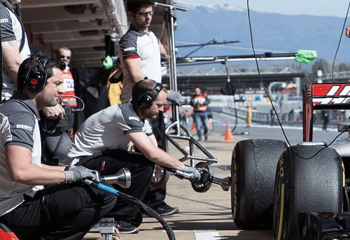 Team of mechanics at a pit stop on a race track, working on the tires of a race car. Other equipment, track boundaries and mountains can be seen in the background