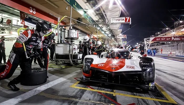 Rennwagen in der Boxengasse bei Nacht, umgeben von Mechanikern in Schutzkleidung. Im Hintergrund Tankstation und Ausrüstung, vorne ein Toyota-Rennfahrzeug bereit für den Einsatz.