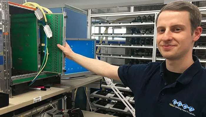 Technischer Arbeitsplatz mit geöffnetem Servergehäuse, Kabeln und grüner Platine. Im Hintergrund sind Regale mit weiteren Geräten und ein Monitor sichtbar.“