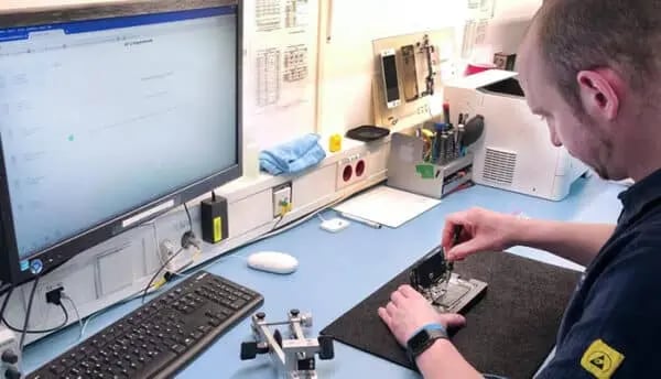 Arbeitsplatz mit Computerbildschirm, Tastatur und Werkzeugen. Eine Person arbeitet an einem kleinen elektronischen Gerät auf einer Matte, umgeben von technischen Komponenten.