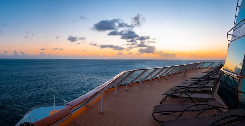 Deck eines Kreuzfahrtschiffs bei Sonnenuntergang mit Blick auf das offene Meer. Mehrere Liegestühle sind entlang der Reling angeordnet, der Himmel zeigt orange und blaue Farbtöne.
