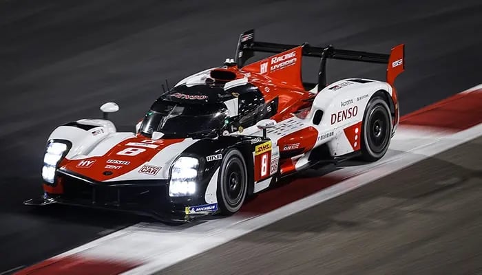 Toyota-Rennwagen mit rotem und weißem Design fährt auf einer beleuchteten Rennstrecke bei Nacht. Sichtbar sind Sponsorenlogos wie Denso und Michelin.